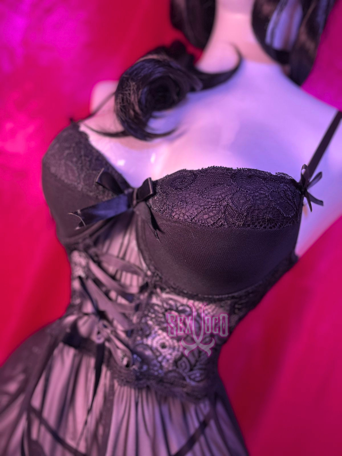 Baby doll negro de encaje con transparencias y detalles de moños en maniquí sobre fondo rosa.